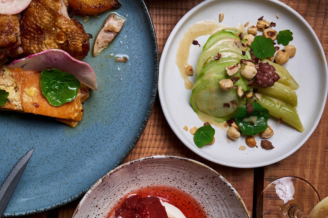 Tre piatti di alta cucina presentati su un tavolo di legno: a sinistra un piatto blu con pesce affumicato e verdure, al centro un piatto bianco con avocado affettato, germogli e nocciole, in basso un piatto grigio con salsa rossa e panna acida. Presentazi