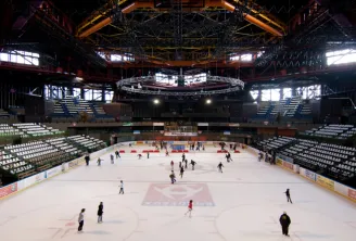 Vista interna della Pista di Ghiaccio Mériadeck a Bordeaux con numerosi pattinatori sul ghiaccio, spalti vuoti e struttura metallica del tetto con illuminazione arancione e gialla.