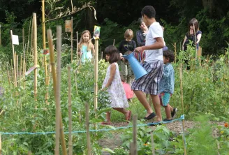 Un gruppo di bambini in un orto del Parco Rivière a Bordeaux, dove un giovane adulto mostra loro come innaffiare le piante con un annaffiatoio blu.