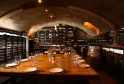 Cantina sotterranea con volta in muratura, tavolo di degustazione in legno massiccio, illuminazione ambientale calda e scaffali pieni di bottiglie di vino di Bordeaux