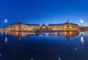 Vista notturna della Place de la Bourse a Bordeaux con edifici storici illuminati riflessi nello specchio d'acqua al tramonto.