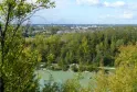 Vista panoramica del Parco dell'Ermitage Sainte-Catherine a Bordeaux con un lago in primo piano, bosco di pioppi e skyline cittadino sotto un cielo nuvoloso