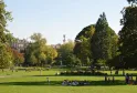 Vista del Giardino Pubblico di Bordeaux con prati verdi, alberi maestosi, visitatori rilassati, un monumento con statua sullo sfondo e edifici storici sotto cielo sereno.