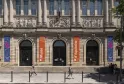 Facciata del Museo d'Aquitania a Bordeaux con colonne imponenti, rilievi scolpiti, striscioni arancioni e blu, tre porte ad arco e visitatori sulla piazza.